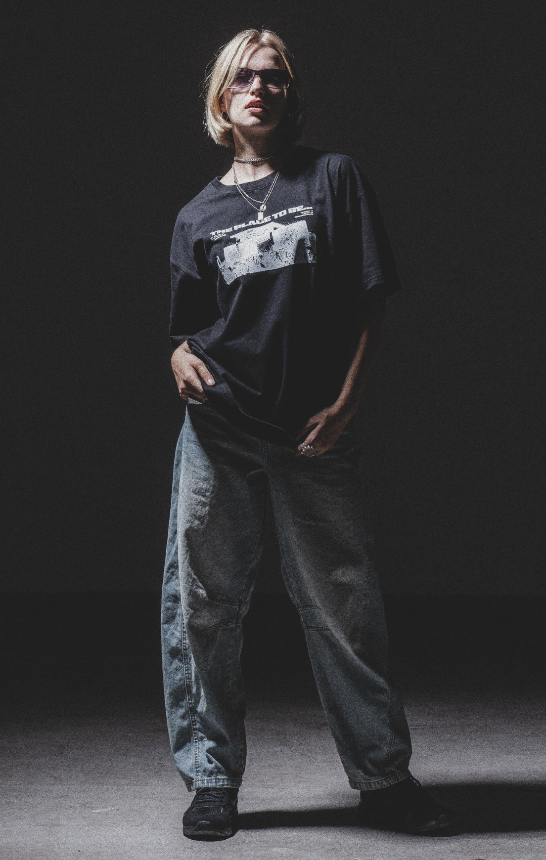 A person with short blond hair wearing dark sunglasses, a black graphic T-shirt, loose-fitting jeans, and black shoes stands confidently in a dimly lit space with a dark background.