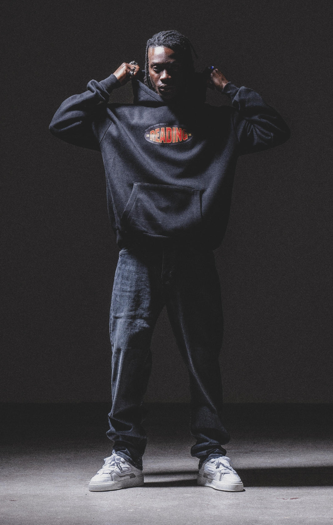 A person stands in a dark room wearing a black hoodie with the word READING in red and orange letters, dark jeans, and white sneakers, holding the hood with both hands.